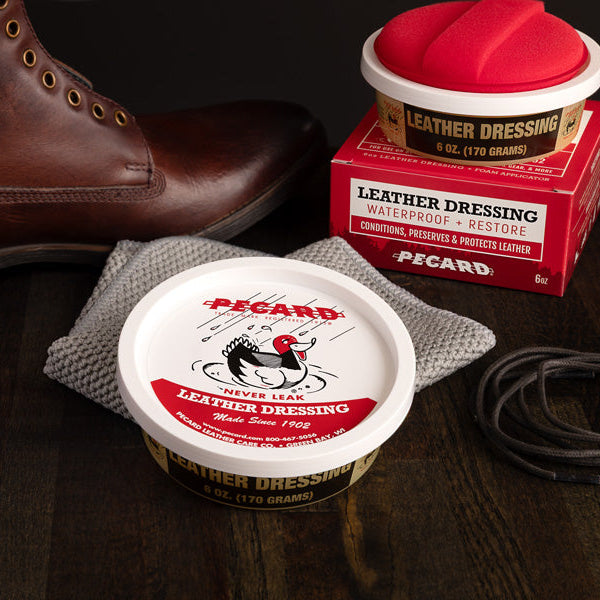 Brown leather boot next to a container of Pecard leather dressing on a wooden surface.