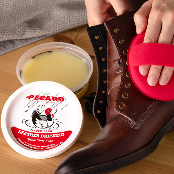 Person applying Pecard leather dressing to a brown boot with a red applicator, next to a container of leather dressing.