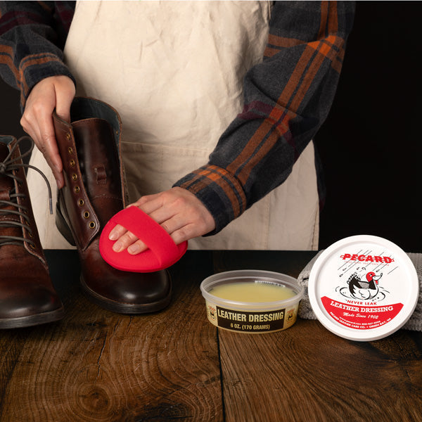 Person cleaning leather boots with a red sponge, surrounded by Pecard leather care products on a wooden surface.